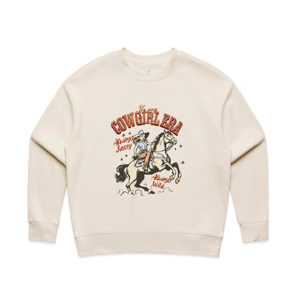 A close-up of a cream-colored t-shirt featuring a vintage-style western graphic. The graphic shows a cowboy riding a rearing horse, with retro text reading 'In my COWGIRL ERA', 'Always Sassy', and 'Always Wild' in reddish-brown and black.