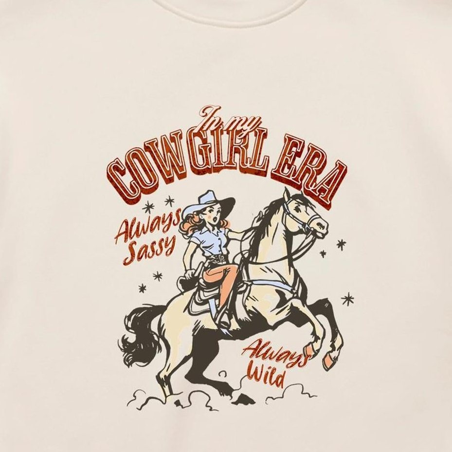 A close-up of a cream-colored t-shirt featuring a vintage-style western graphic. The graphic shows a cowboy riding a rearing horse, with retro text reading 'In my COWGIRL ERA', 'Always Sassy', and 'Always Wild' in reddish-brown and black.