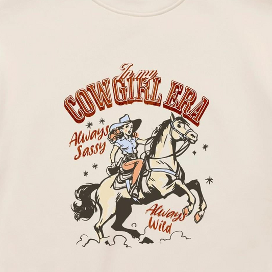 A close-up of a cream-colored t-shirt featuring a vintage-style western graphic. The graphic shows a cowboy riding a rearing horse, with retro text reading 'In my COWGIRL ERA', 'Always Sassy', and 'Always Wild' in reddish-brown and black.