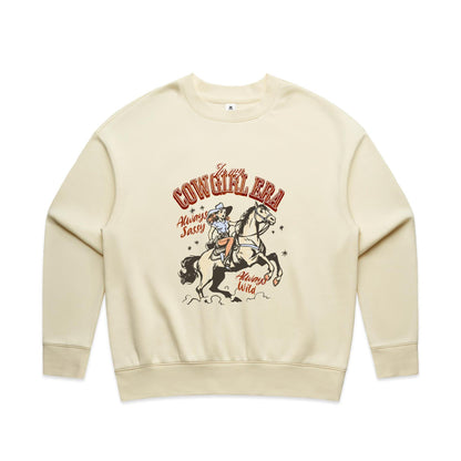 A close-up of a cream-colored t-shirt featuring a vintage-style western graphic. The graphic shows a cowboy riding a rearing horse, with retro text reading 'In my COWGIRL ERA', 'Always Sassy', and 'Always Wild' in reddish-brown and black.