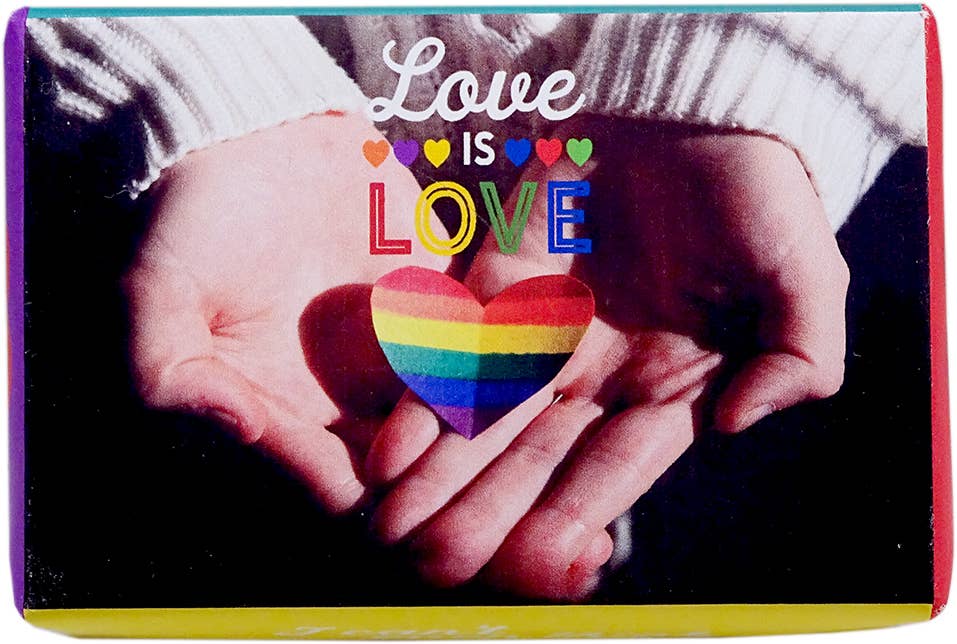 A square greeting card or print featuring a close-up of two hands holding a rainbow-striped heart, with the words 'Love IS LOVE' printed above. The card has a colorful border and is photographed against a dark background.