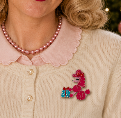 A close-up lifestyle shot of a woman wearing a pink poodle pin with blue and white polka-dot gift boxes, styled with a pink collar sweater and pearl necklace.