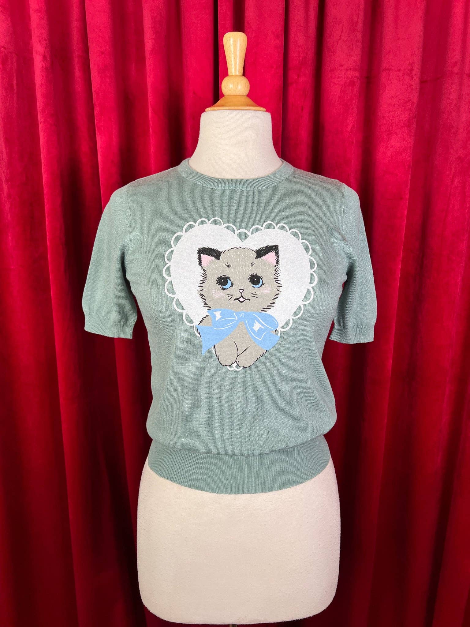 Green sweater with a cat design on a mannequin against a red curtain background