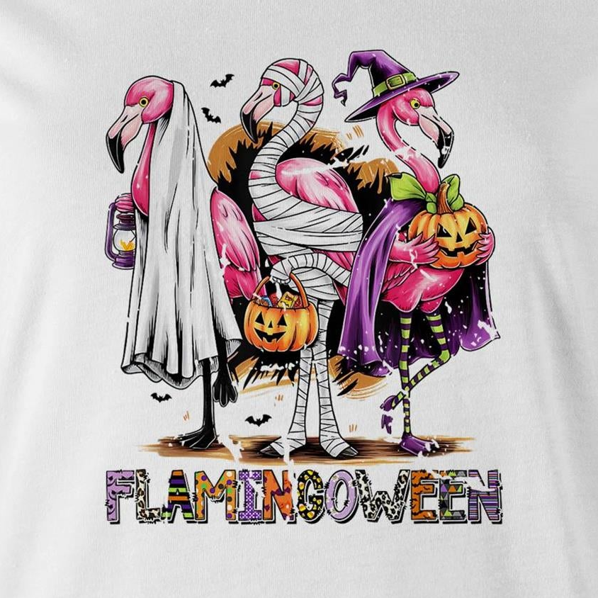 A white t-shirt featuring a colorful graphic of three cartoon flamingos dressed in Halloween costumes. One flamingo wears a white ghost and lantern, another is wrapped in bandages and holds a pumpkin bucket, and the third wears a purple witch hat and cape while holding a jack-o'-lantern.
