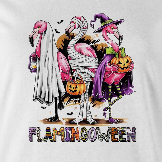A white t-shirt featuring a colorful graphic of three cartoon flamingos dressed in Halloween costumes. One flamingo wears a white ghost and lantern, another is wrapped in bandages and holds a pumpkin bucket, and the third wears a purple witch hat and cape while holding a jack-o'-lantern.