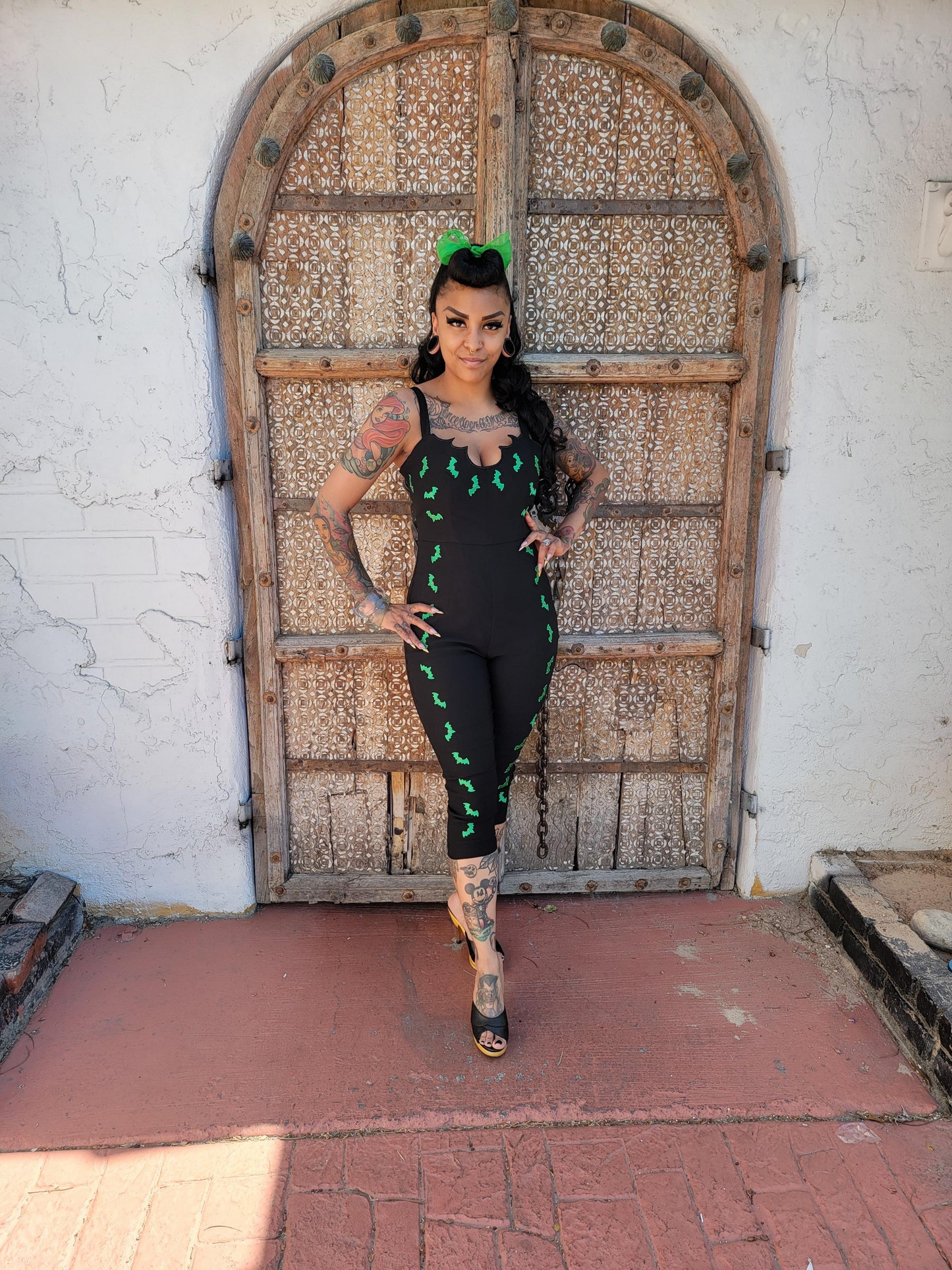 A woman stands in front of a large rustic wooden door wearing a black sleeveless jumpsuit with green embroidered details and a green headband.