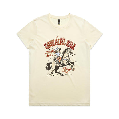 A white graphic t-shirt featuring a retro western illustration of a cowgirl riding a horse. The text reads 'In my COWGIRL ERA' in arched red letters, 'Always Sassy' in red script, and 'Always Wild' in red script, with the cowgirl wearing a cowboy hat and boots while riding a rearing horse.