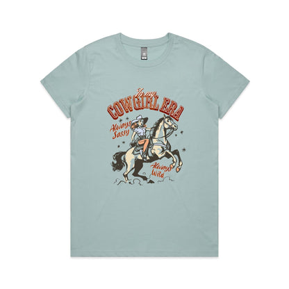 A white graphic t-shirt featuring a retro western illustration of a cowgirl riding a horse. The text reads 'In my COWGIRL ERA' in arched red letters, 'Always Sassy' in red script, and 'Always Wild' in red script, with the cowgirl wearing a cowboy hat and boots while riding a rearing horse.