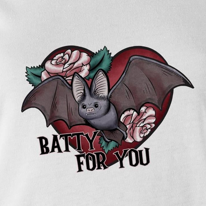 A close-up of a white fabric surface featuring a graphic print of a cute gray bat in front of a red heart, decorated with pink roses and green leaves, with the text 'BATTY FOR YOU' written below in black.