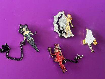 Four Halloween-themed enamel pins arranged on a solid purple background: a black cat with a chain, a silver spider-themed fairy, a red witch fairy, and a white ghost with a gold pumpkin.