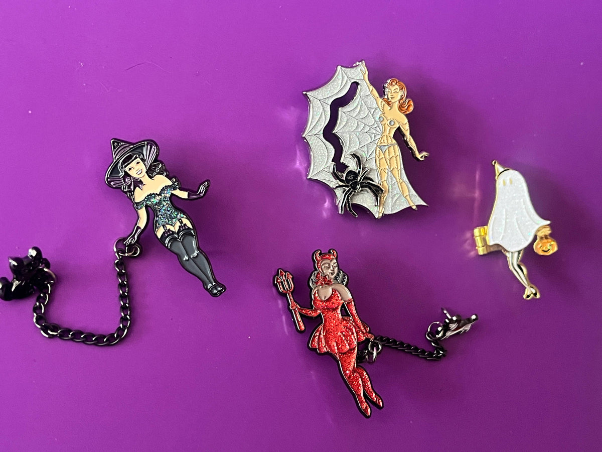 Four Halloween-themed enamel pins arranged on a solid purple background: a black cat with a chain, a silver spider-themed fairy, a red witch fairy, and a white ghost with a gold pumpkin.