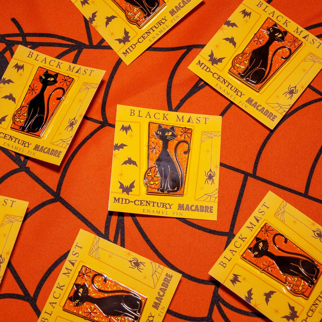 Several yellow Halloween enamel pins featuring a black cat in a lantern, labeled 'BLACK MAST MID-CENTURY MACABRE', arranged on an orange background with a black spiderweb pattern.