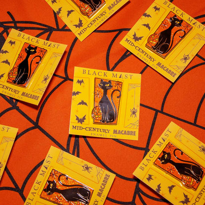 Several yellow Halloween enamel pins featuring a black cat in a lantern, labeled 'BLACK MAST MID-CENTURY MACABRE', arranged on an orange background with a black spiderweb pattern.