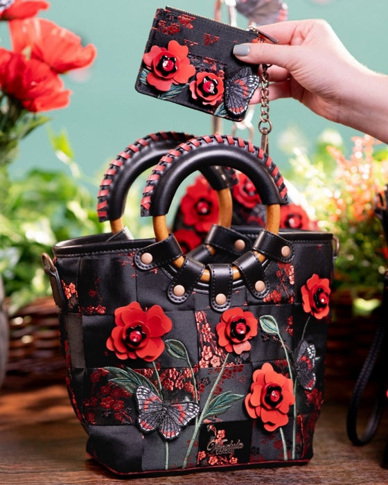 Black handbag with red floral designs held by a hand against a floral background