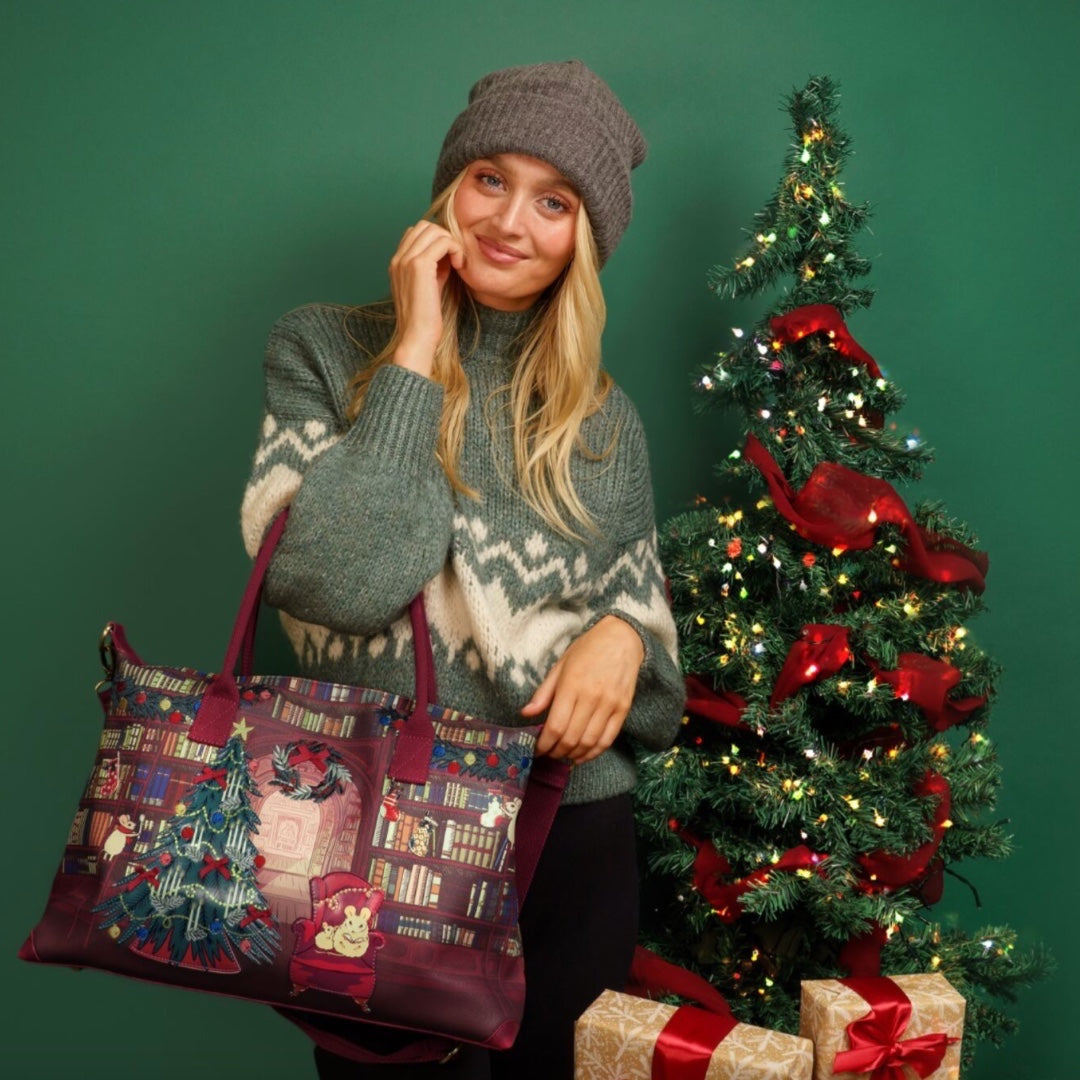 Woman holding a decorative bag with a Christmas tree and presents design, standing next to a decorated Christmas tree.