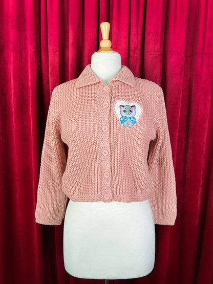 Pink knitted cardigan with a cat patch on a mannequin against a red curtain background
