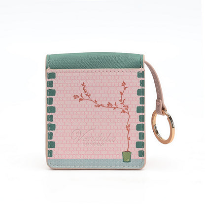 A small square card holder wallet by Vendimia London, featuring a pink patterned front with a botanical branch illustration, a teal top panel, a pale blue bottom border, and a tan wrist strap with a gold-tone ring on a white background.