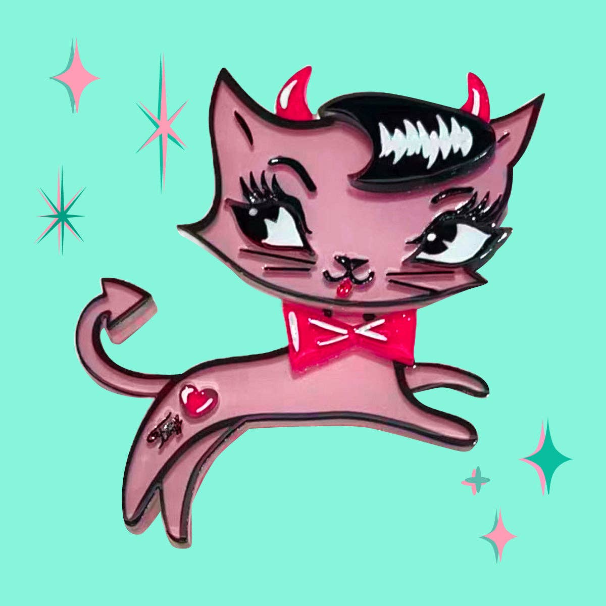 A cartoon-style pink cat pin or badge with red devil horns, a red bow tie, and a small red heart on its tail, shown on a light aqua background with decorative starburst accents.
