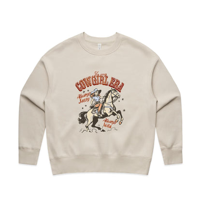 A close-up of a cream-colored t-shirt featuring a vintage-style western graphic. The graphic shows a cowboy riding a rearing horse, with retro text reading 'In my COWGIRL ERA', 'Always Sassy', and 'Always Wild' in reddish-brown and black.
