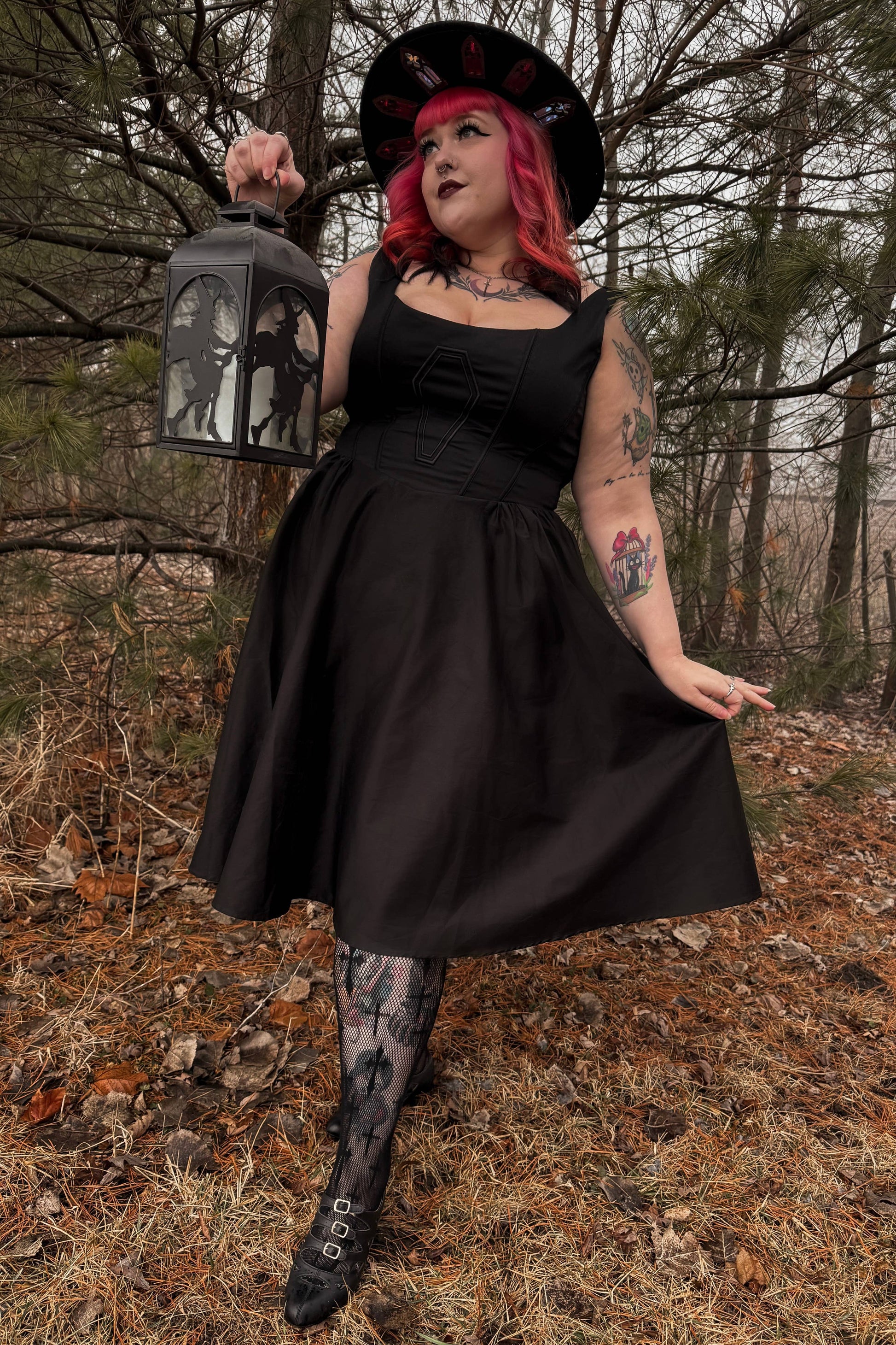 Person in a black dress and hat holding a lantern in a forest setting