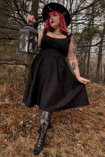 Person in a black dress and hat holding a lantern in a forest setting