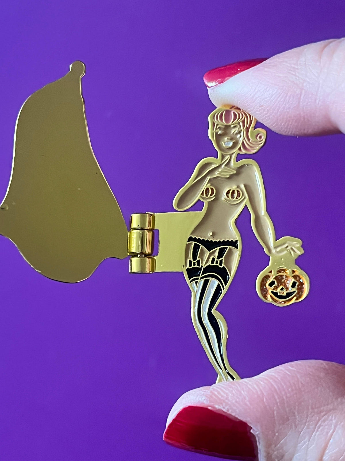 A gold-colored metal pin shaped like a pin-up woman holding a jack-o'-lantern, held by fingers with red nail polish against a solid purple background.