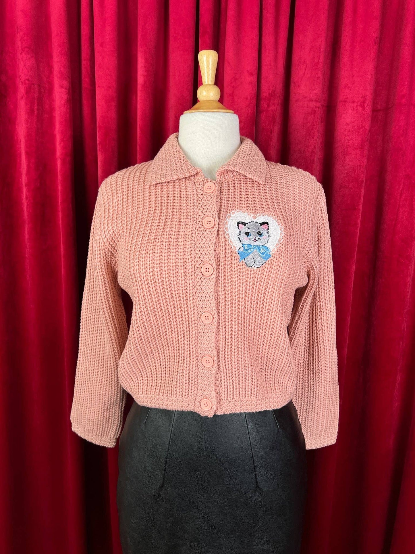 Pink knitted cardigan with a cat patch on a mannequin against a red curtain background