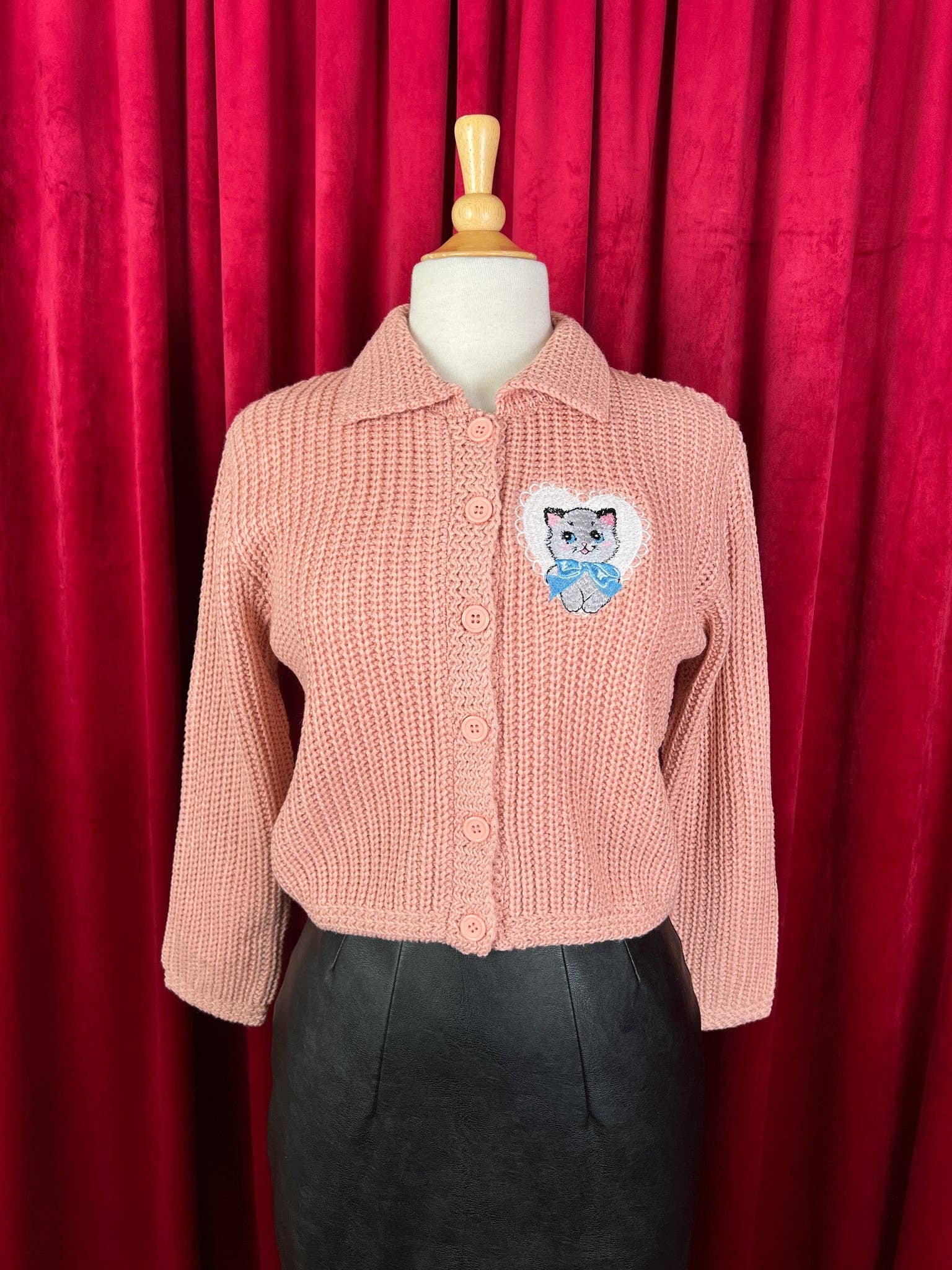 Pink knitted cardigan with a cat patch on a mannequin against a red curtain background