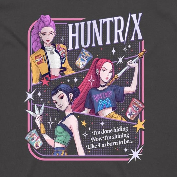 A dark charcoal gray t-shirt featuring a colorful graphic print of three anime-style female characters with pink, purple, and black hair, holding snacks and a sword, with the text 'HUNTR/X' and 'I'm done hiding Now I'm shining Like I'm born to be.