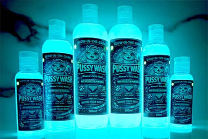 Six glow-in-the-dark bottles of Pussy Wash personal care products arranged in a row against a dark, black background with a faint silhouette.