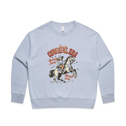 A close-up of a cream-colored t-shirt featuring a vintage-style western graphic. The graphic shows a cowboy riding a rearing horse, with retro text reading 'In my COWGIRL ERA', 'Always Sassy', and 'Always Wild' in reddish-brown and black.