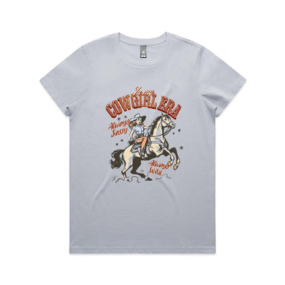 A white graphic t-shirt featuring a retro western illustration of a cowgirl riding a horse. The text reads 'In my COWGIRL ERA' in arched red letters, 'Always Sassy' in red script, and 'Always Wild' in red script, with the cowgirl wearing a cowboy hat and boots while riding a rearing horse.