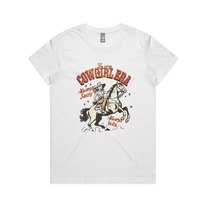 A white graphic t-shirt featuring a retro western illustration of a cowgirl riding a horse. The text reads 'In my COWGIRL ERA' in arched red letters, 'Always Sassy' in red script, and 'Always Wild' in red script, with the cowgirl wearing a cowboy hat and boots while riding a rearing horse.