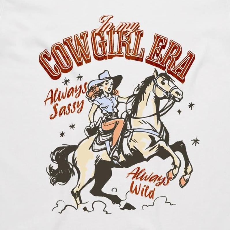 A white graphic t-shirt featuring a retro western illustration of a cowgirl riding a horse. The text reads 'In my COWGIRL ERA' in arched red letters, 'Always Sassy' in red script, and 'Always Wild' in red script, with the cowgirl wearing a cowboy hat and boots while riding a rearing horse.