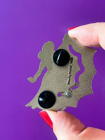 A close-up of a hand with red nail polish holding a silver bat-shaped pin with black oval studs against a solid purple background.