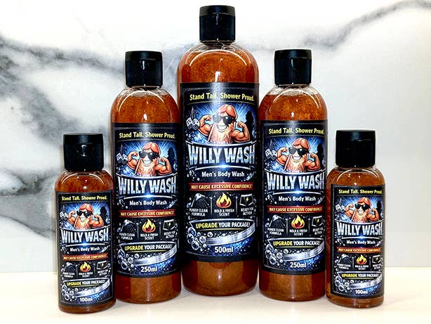 Five bottles of Willy Wash Men's Body Wash in various 250ml and 500ml sizes, arranged in a row against a white marble background. The bottles are filled with amber-colored liquid and feature black caps and colorful labels with a cartoon graphic of a man in a cowboy hat.