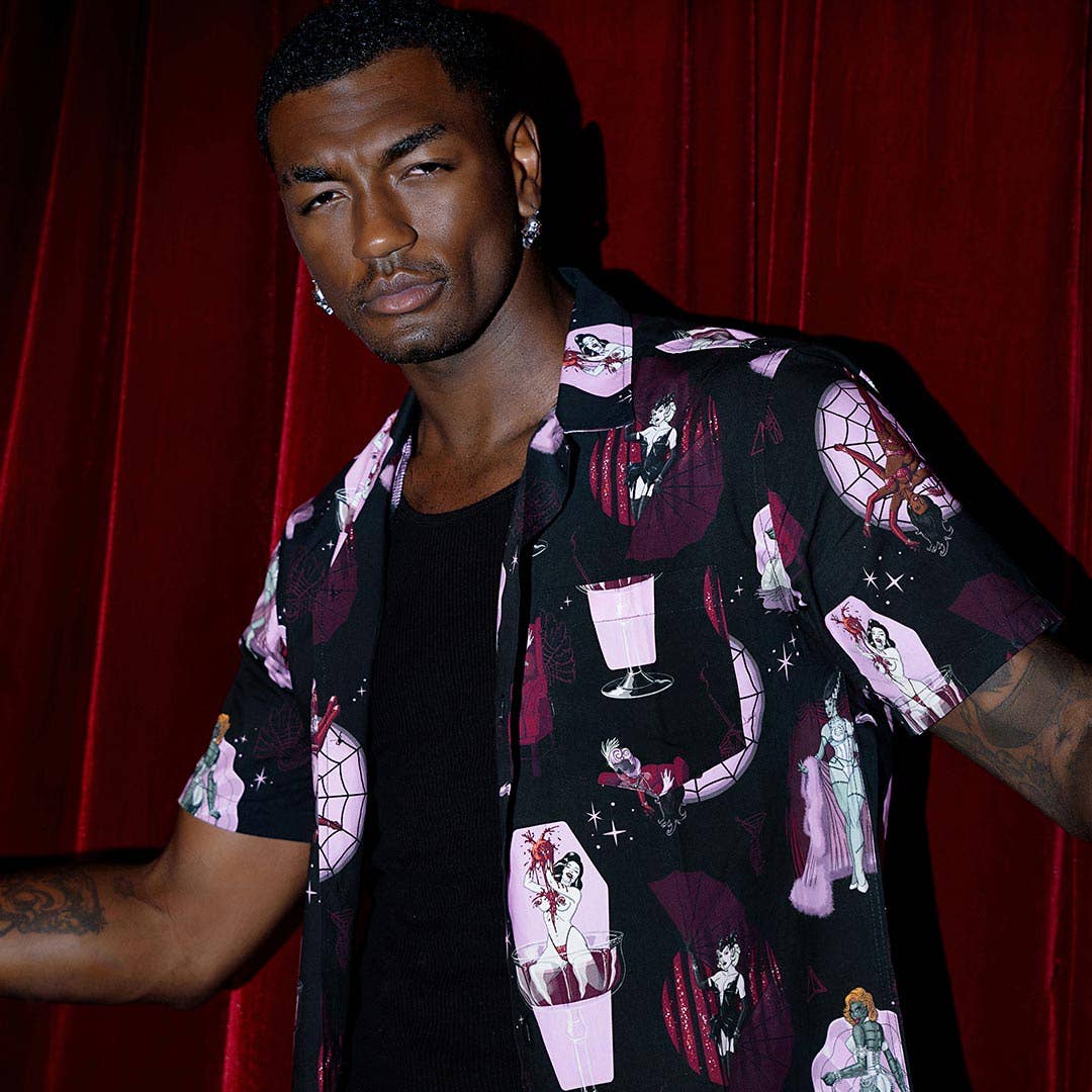 A man wearing a black short-sleeve button-up shirt with a gothic and horror-themed print featuring skulls, caskets, and a cocktail glass, posed against a red curtain background.