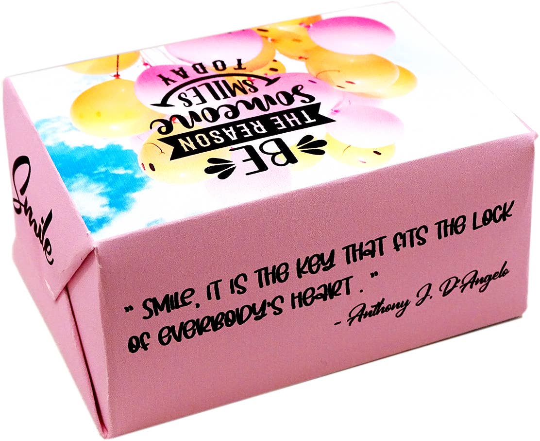 A pink cardboard gift box with a printed smiley-face balloon graphic on the lid and a black quote on the side. The quote reads, 'smile, it is the key that fits the lock of everybody's heart.