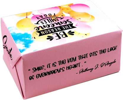 A pink cardboard gift box with a printed smiley-face balloon graphic on the lid and a black quote on the side. The quote reads, 'smile, it is the key that fits the lock of everybody's heart.