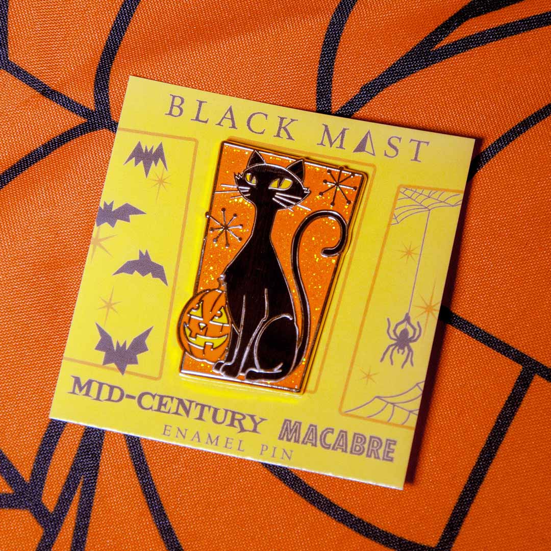 A black cat enamel pin with glowing yellow eyes and a pumpkin, mounted on a yellow backing card labeled 'BLACK MAEST' and 'MID-CENTURY MACABRE ENAMEL PIN,' placed on an orange patterned fabric background.