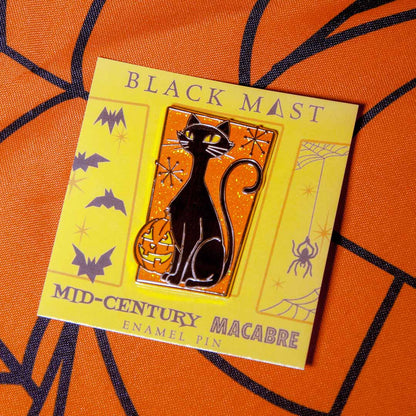 A black cat enamel pin with glowing yellow eyes and a pumpkin, mounted on a yellow backing card labeled 'BLACK MAEST' and 'MID-CENTURY MACABRE ENAMEL PIN,' placed on an orange patterned fabric background.