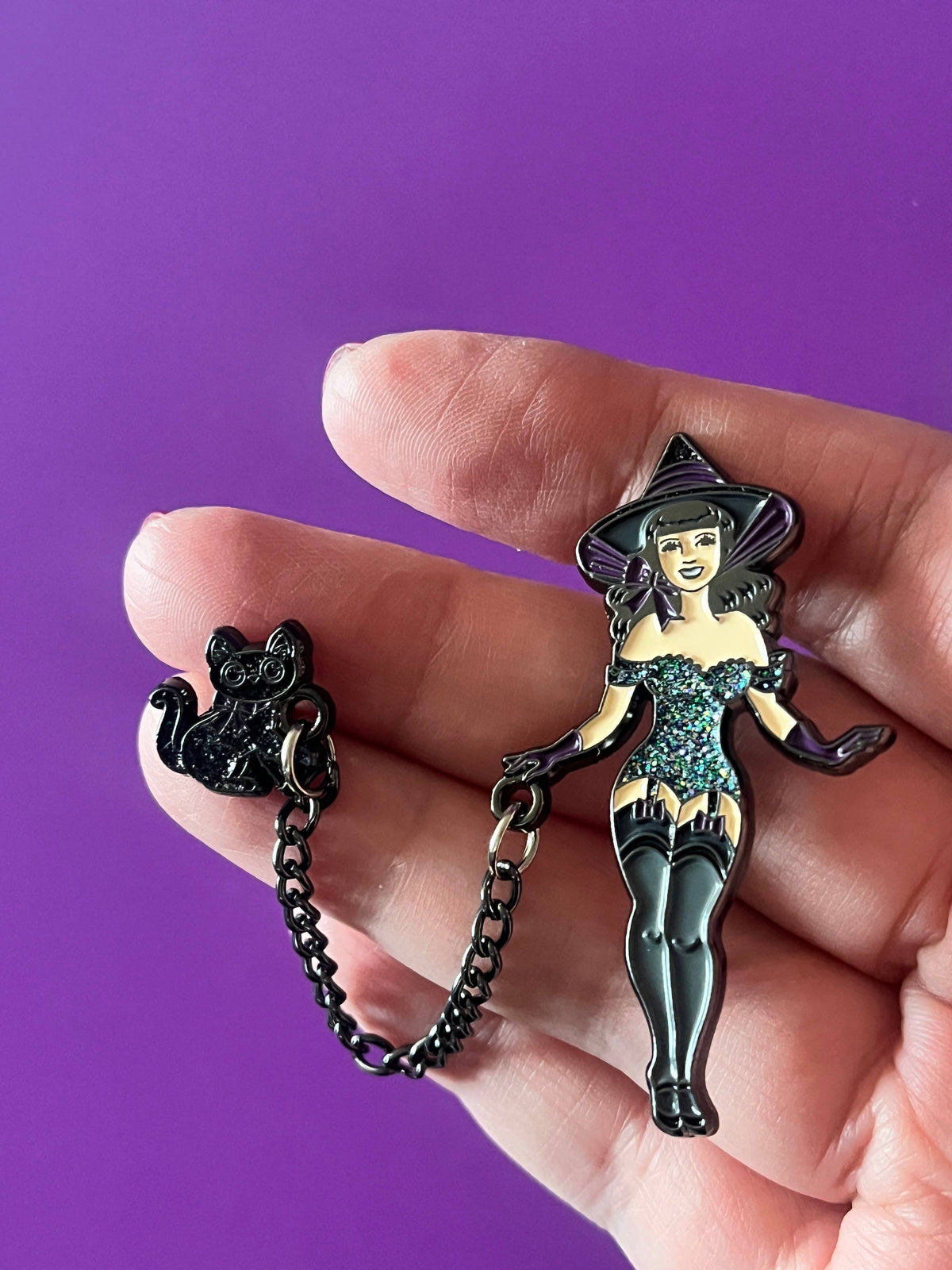 A hand holds a black witch-themed enamel pin set against a solid purple background. The set includes a witch pin in a sparkling teal dress and black hat, attached to a small black cat charm by a short black chain.