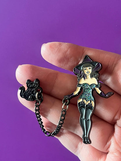 A hand holds a black witch-themed enamel pin set against a solid purple background. The set includes a witch pin in a sparkling teal dress and black hat, attached to a small black cat charm by a short black chain.