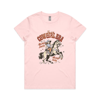 A white graphic t-shirt featuring a retro western illustration of a cowgirl riding a horse. The text reads 'In my COWGIRL ERA' in arched red letters, 'Always Sassy' in red script, and 'Always Wild' in red script, with the cowgirl wearing a cowboy hat and boots while riding a rearing horse.