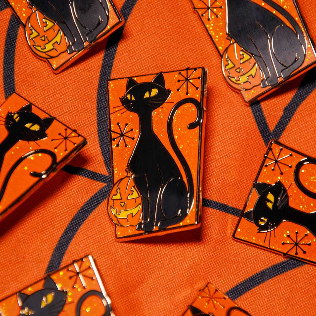 A close-up of several Halloween-themed enamel pins featuring a black cat and jack-o'-lantern on an orange glittery background, arranged on an orange patterned fabric.