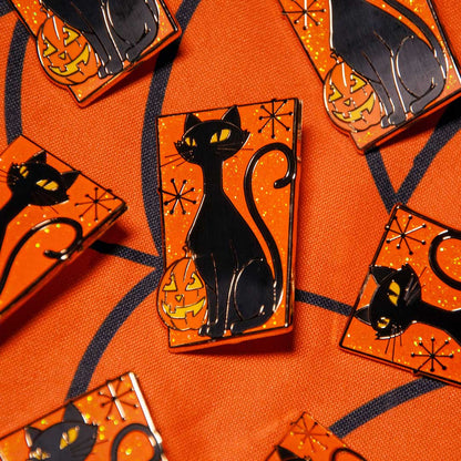 A close-up of several Halloween-themed enamel pins featuring a black cat and jack-o'-lantern on an orange glittery background, arranged on an orange patterned fabric.
