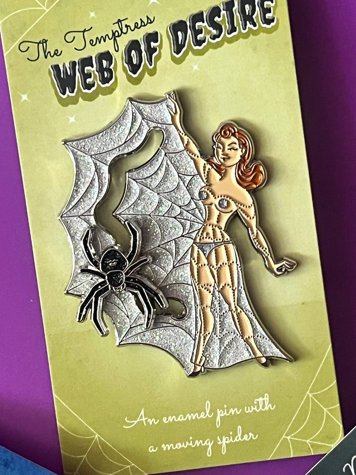 The Temptress Web of Desire enamel pin on its original light green card packaging, featuring a silver spider, a woman's skeleton body, and a large web design, set against a purple background.
