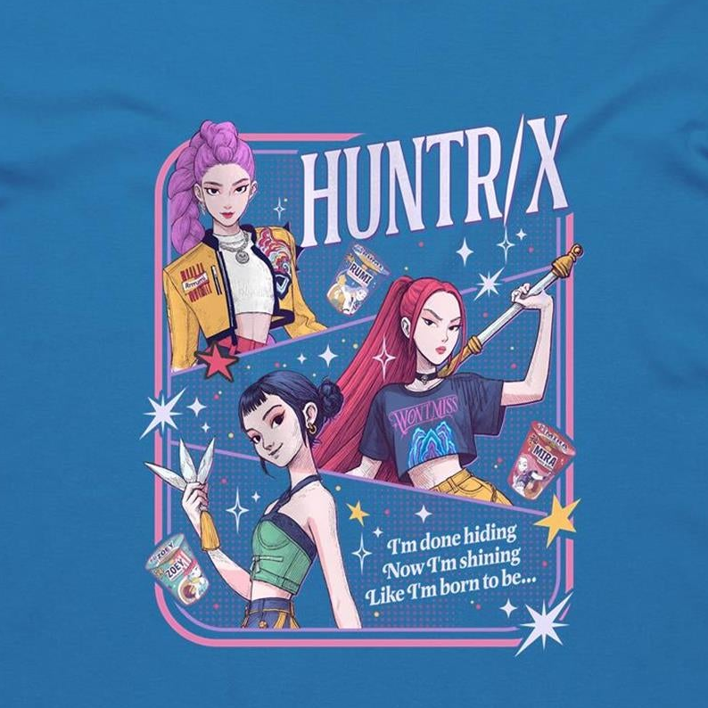 A blue graphic t-shirt featuring a colorful retro-style illustration of three anime characters with pink, purple, and red hair, alongside the text 'HUNTR/X' and a quote. The graphic is framed by a pink and blue border with starburst accents.