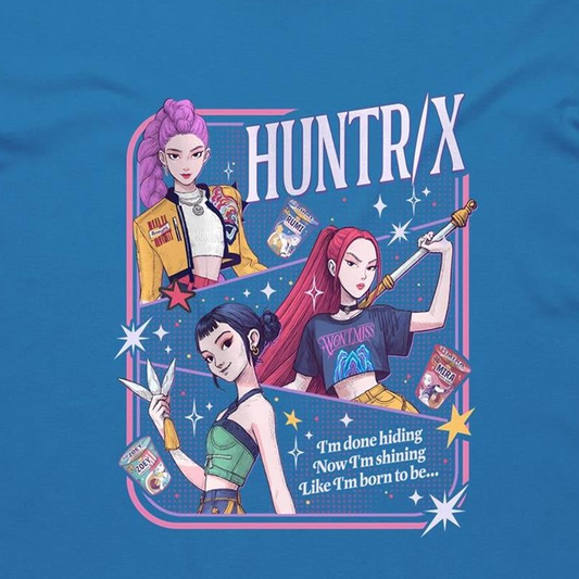 A blue graphic t-shirt featuring a colorful retro-style illustration of three anime characters with pink, purple, and red hair, alongside the text 'HUNTR/X' and a quote. The graphic is framed by a pink and blue border with starburst accents.