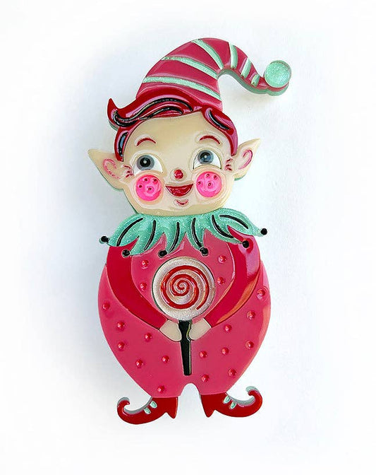 A whimsical enamel pin shaped like a strawberry with a small elf character on top, holding a lollipop. The pin features a vibrant pink strawberry body with red seeds, a green collar, and a red-and-green striped hat, set against a plain white background.